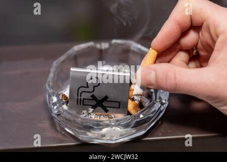 Bavaria, Germany - January 6, 2024: Smoke-free concept, a man puts out a burning cigarette in an ashtray with a no smoking sign *** Rauchfrei Konzept, ein Mann drückt eine brennende Zigarette in einem Aschenbecher mit einem Rauchverbot Schild aus Stock Photo