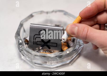 Bavaria, Germany - January 6, 2024: Smoke-free concept, a man puts out a burning cigarette in an ashtray with a no smoking sign *** Rauchfrei Konzept, ein Mann drückt eine brennende Zigarette in einem Aschenbecher mit einem Rauchverbot Schild aus Stock Photo