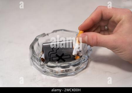 Bavaria, Germany - January 6, 2024: Smoke-free concept, a man puts out a burning cigarette in an ashtray with a no smoking sign *** Rauchfrei Konzept, ein Mann drückt eine brennende Zigarette in einem Aschenbecher mit einem Rauchverbot Schild aus Stock Photo