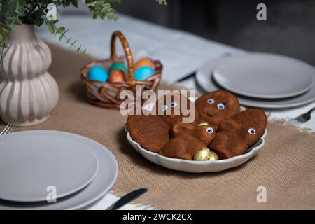 Easter table setting with empty gray plates, multi colored eggs in wicker basket and vase with green plant in on tablecloth Stock Photo