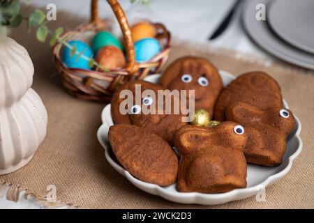 Close-up of funny muffins shaped as bunnies and eggs with eyes, multi colored Easter eggs in wicker basket on served table Stock Photo