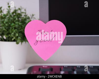 A pink heart shaped paper note with words Feel Good on it sticked on to a monitor at an office workplace. Stock Photo