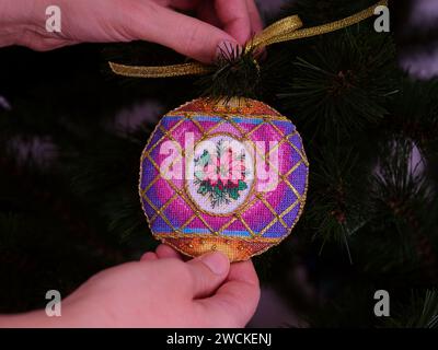 A woman hanging a purple cross stitched Christmas Timeless Elegance ornament on to a Christmas tree. This Christmas ornament with poinsettia embroider Stock Photo