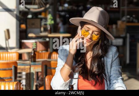 Woman in sunglasses and hat talking on the phone while sitting in a coffee shop. Technology concept. Stock Photo