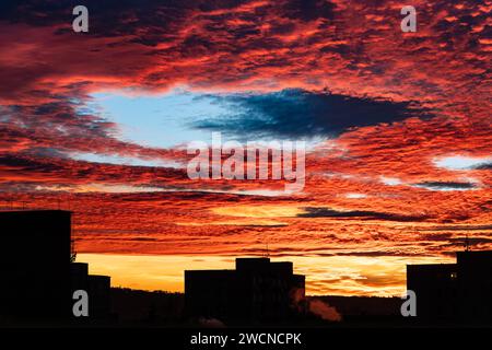 Beautiful sunset with fallstreak hole in red fiery sky and a buildings silhouette. Majestic orange clouds with large blue gap at scenic winter dusk. Stock Photo