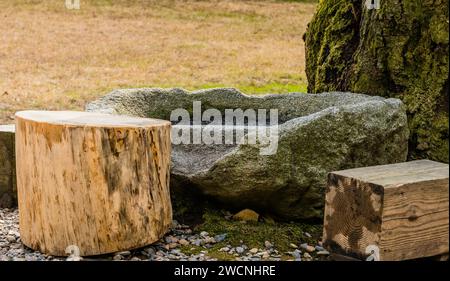 Man made concrete stool and three wooden stools on ground covered with small rocks next to tree covered in green moss Stock Photo