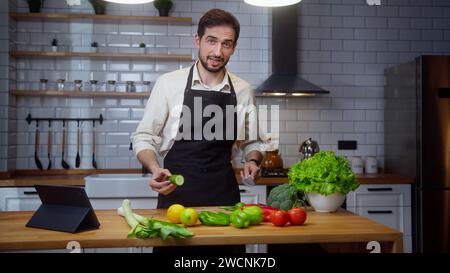 Bearded young vlogger man chef in an apron recording video cooking lesson about vegetarian meal in modern kitchen Stock Photo