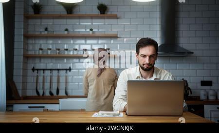 Young man is working on his laptop in the kitchen at home while his wife working in kitchen Stock Photo