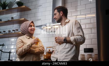 Young couple drinking orange juice, having conversation at home kitchen in morning while standing Stock Photo