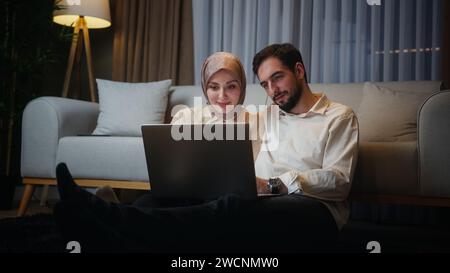 Young married couple leaning against the sofa sitting on floor at home at night watching show or film, spending great time Stock Photo