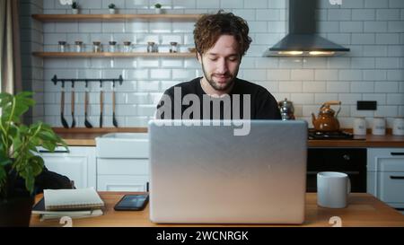 Handsome man sitting in the kitchen using laptop enjoying surfing internet, using social media, chatting with friends, leaning back and smiling Stock Photo