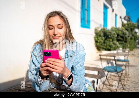 Beautiful blond woman using smartphone at outdoor cafe table. Happy female tourist in denim shirt sitting near white building. Lady enjoys her vacation in typical Portugal city. Stock Photo