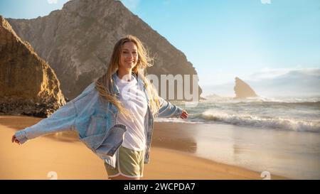 Portrait of smiling blond woman standing arms outstretched on sandy beach. Beautiful female in denim jacket and shorts standing near big rock and sea waves. Lady enjoys summer vacation. Stock Photo