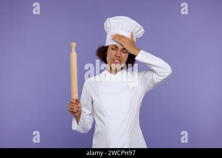 Frustrated female chef in uniform holding rolling pin on purple background Stock Photo