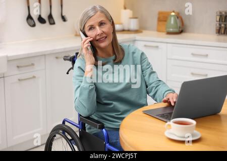 Woman in wheelchair talking on smartphone while using laptop at home Stock Photo