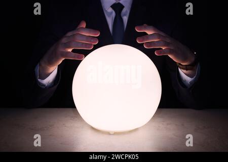 Businessman using glowing crystal ball to predict future at table in darkness, closeup. Fortune telling Stock Photo