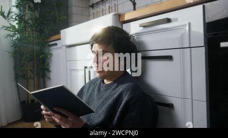 Young man sitting on the floor in the kitchen looking notebook in his hand, reading, smiling Stock Photo