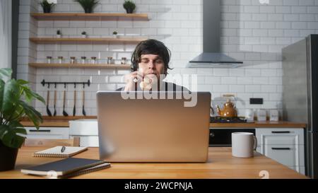 Focused male sitting in kitchen at home wear wireless headset with microphone having video conference calling on laptop computer talk by webcam Stock Photo