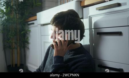 Young man sitting on the floor in kitchen talking by the cell phone, having informal pleasant conversation, share news, enjoy remote communication Stock Photo