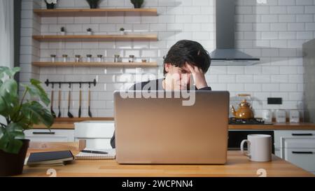 Young man sitting in the kitchen counter headache while using laptop. Tired young man get having pain from a headache or migraine Stock Photo