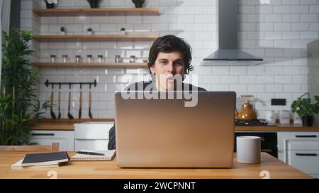 Surprised man sitting in the kitchen having video call conversation through using laptop computer. Share news, talk to family, enjoy webchat virtual m Stock Photo