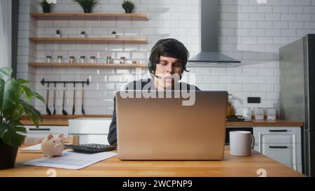 Focused male sitting in kitchen at home wear wireless headset with microphone having video conference calling on laptop computer talk by webcam, learn Stock Photo