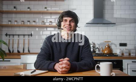 Young man wears wireless headset with microphone talking to camera making conducting conference business call in kitchen. Having video call zoom meeti Stock Photo