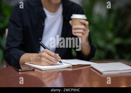Close-up image of a young Asian man writing something in his book and sipping coffee at a table in a coffee shop. working remotely, digital nomad, fre Stock Photo