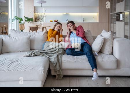 Scared man woman closing faces with hands watching horror movies eating popcorn. Home cinema Stock Photo