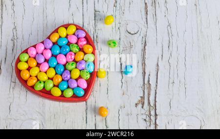 Heart-shaped box filled with colorful Easter candy eggs on white rustic wooden background, copy space Stock Photo