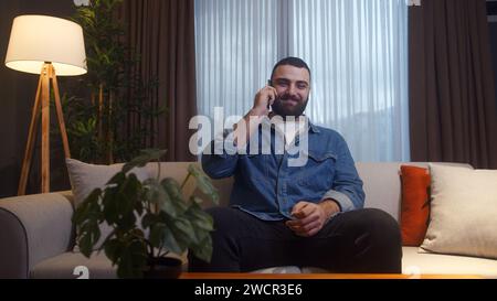 Smiling young adult bearded man sitting on sofa while talking by the cell phone at home, having informal pleasant conversation, share news spend time Stock Photo