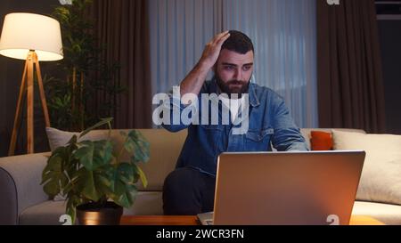 Young adult man sitting on sofa at home while using laptop having anxiety and stress. Upset young man reacting to loss, bad news on laptop Stock Photo