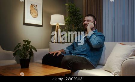Smiling young adult bearded man sitting on sofa while talking by the cell phone at home, having informal pleasant conversation, share news Stock Photo