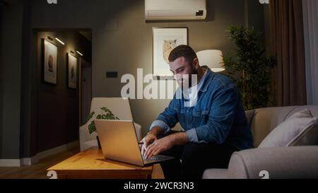 Bearded young freelancer working from home with using laptop while sitting on sofa at home. Young man looking at screen typing on computer getting job Stock Photo