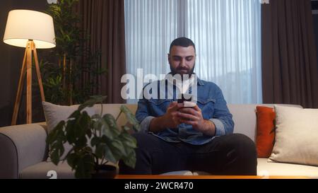 Handsome young adult man sitting on couch at home typing text message by smartphone. Male person relaxing at home. Communicating with family and frien Stock Photo
