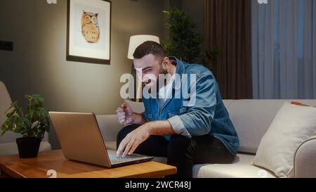 Young man sitting on sofa while using laptop checking messages read notice receive good news on email celebrate great opportunity feels happy Stock Photo