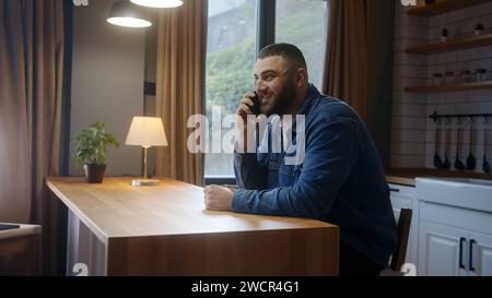 Smiling young man sitting against the kitchen counter talking by the cell phone at home, having informal pleasant conversation, share news, enjoy remo Stock Photo