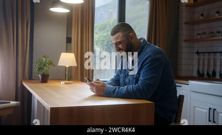 Bearded happy young man sitting against the kitchen counter typing text message by smartphone. Male person relaxing at home, communicating with family Stock Photo