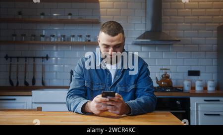 Bearded young adult man sitting against the kitchen counter typing text message by smartphone. Male person relaxing at home, communicating with family Stock Photo