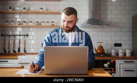 Bearded young adult man wearing in-ear headphones typing on laptop doing research sitting against the kitchen working on laptop Stock Photo