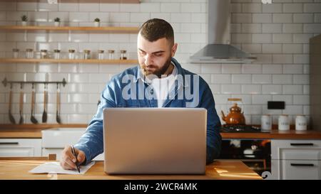 Bearded young adult man wearing in-ear headphones typing on laptop doing research sitting against the kitchen working on laptop Stock Photo