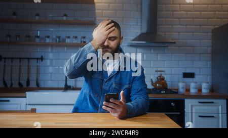 Young adult man sitting against the kitchen counter using smartphone having anxiety and stress. Upset man reacting to bad news Stock Photo