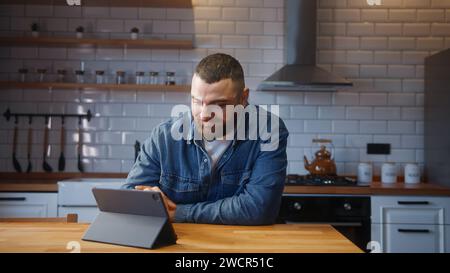 Cozy bearded young adult man sitting against the kitchen counter relaxing and using tablet device surfing internet, using social media or app Stock Photo