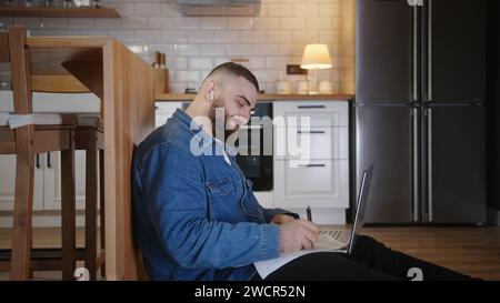 Young man student sitting on the floor leaning against the kitchen counter doing research working on laptop taking notes on paper while listening the Stock Photo