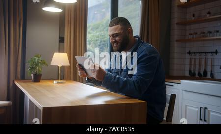 Handsome young adult bearded man playing video game on a tablet in kitchen. Excited concentrated male engrossed in interesting game Stock Photo