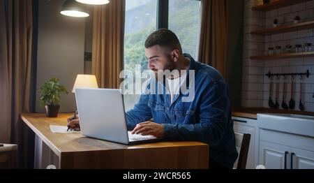 Bearded young adult business man sitting against the kitchen counter typing on keyboard, doing research working on laptop taking notes on paper. Worki Stock Photo