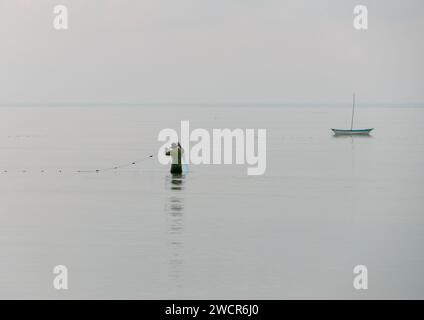 Isolated fisherman stands in shallow sea with long fishing net.  There is a small boat in the background.  The sea is flat calm. Stock Photo