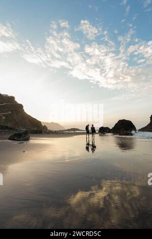 Two silhouetted figures stand near the water edge at Benijo Beach, Tenerife, reflecting on the wet sand during the tranquil sunset Stock Photo