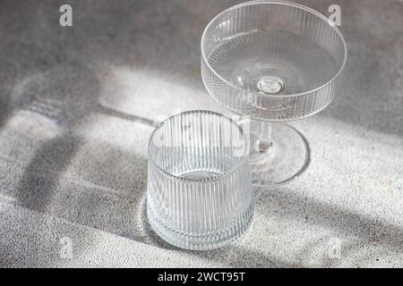 A crystal clear cocktail glass and tumbler sit on a textured surface, their distinct shadows cast by soft, natural light Stock Photo