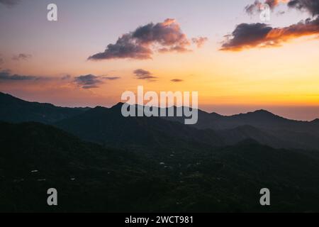 A tranquil sunset over layered mountain peaks with a warm, orange-toned sky and scattered clouds Stock Photo